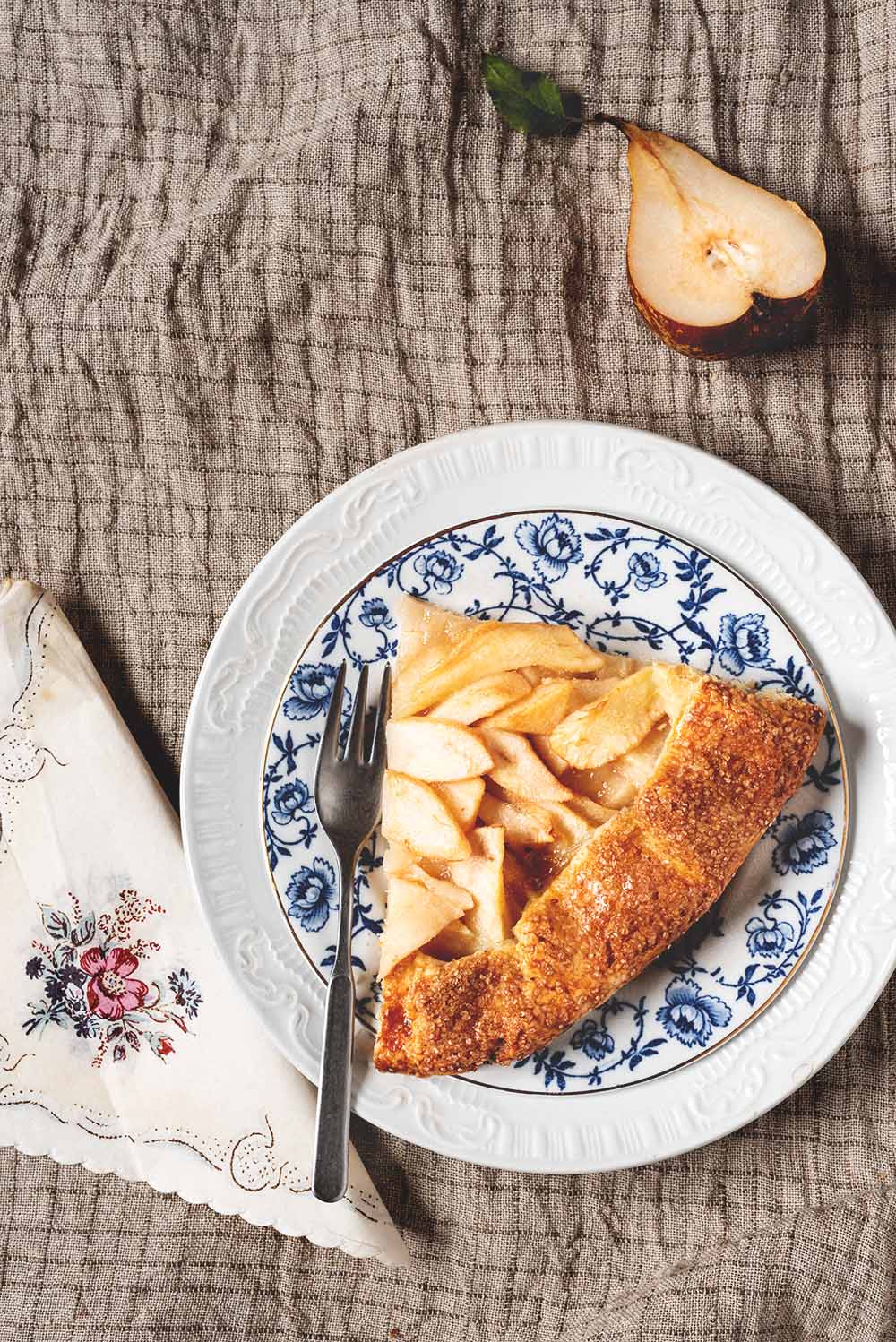 Cómo hacer galette de peras Se ve un mantel gris vintage. En el mantel hay un plato blanco y azul dónde está una ración de galette de peras. Alrededor del postre hay una servilleta de papel vintage y una pera cortada.