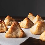 En una mesa de madera hay papel de horno. En el papel hay varios coquitos caseros y esponjosos. Se ven dorados con forma de pirámides.