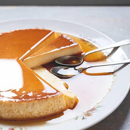 En una mesa de gris hay un plato blanco vintage grande.En el plato está el flan de vainilla al horno ya cortado con su salsa de caramelo. Se ve muy cremosa. En el plato hay dos cucharas.