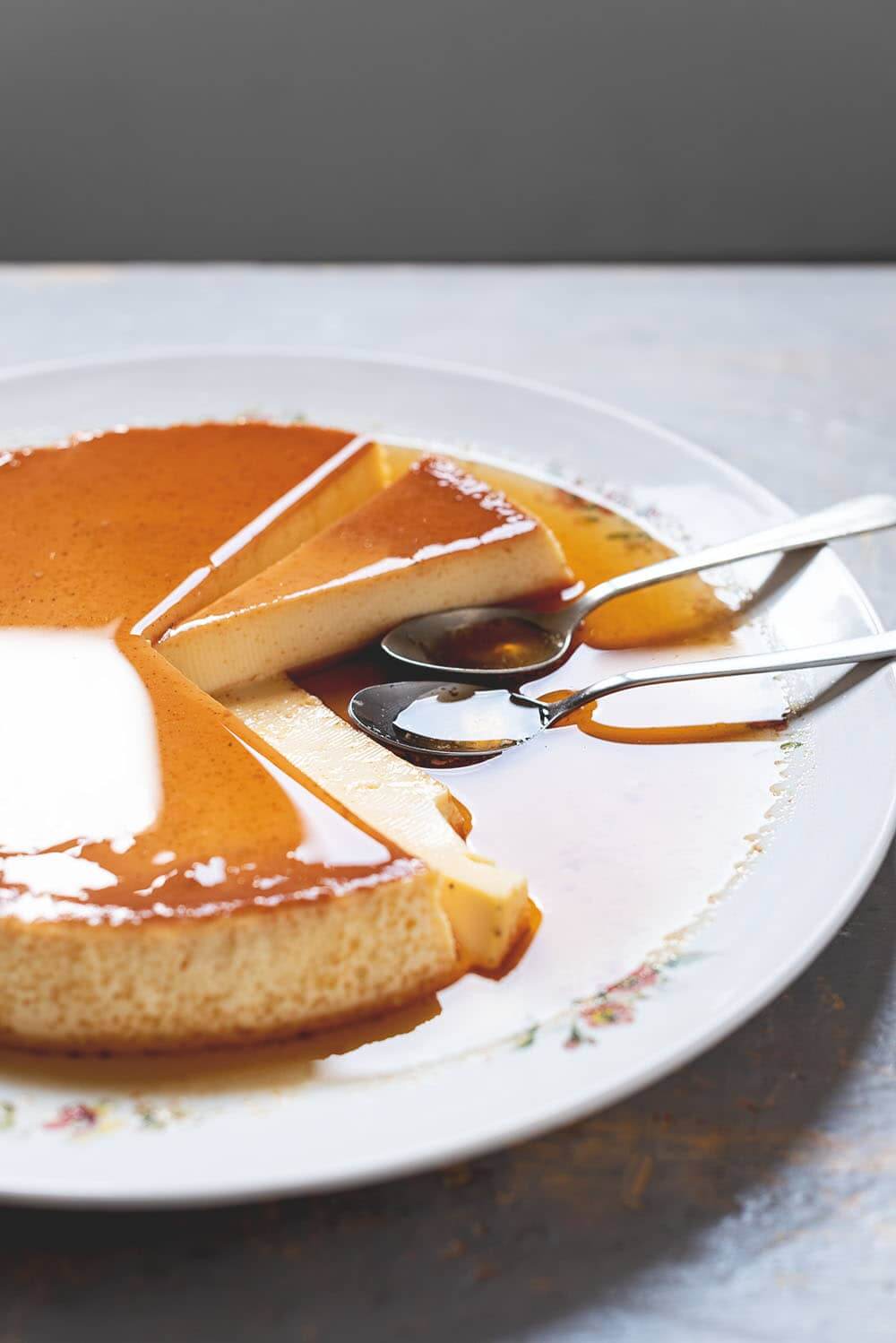 En una mesa de gris hay un plato blanco vintage grande.En el plato está el flan de vainilla al horno ya cortado con su salsa de caramelo. Se ve muy cremosa. En el plato hay dos cucharas.
