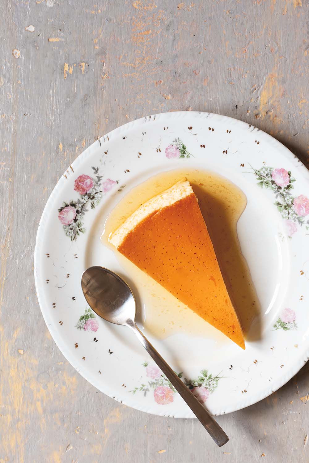 En una mesa de gris hay un plato blanco vintage. En el plato hay una ración de flan de vainilla al horno con su salsa de caramelo. Al lado del flan hay una cuchara.