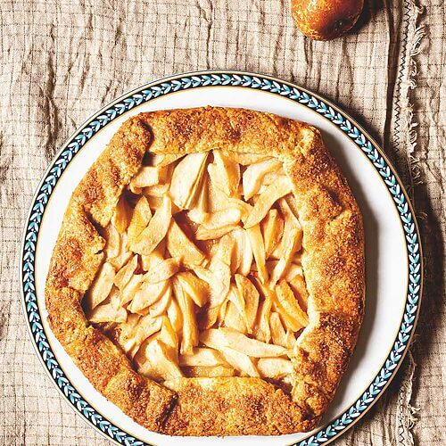 Se ve un mantel gris vintage. En el mantel hay un plato blanco dónde está la galette de peras francesa. Al lado del plato blanco hay una peras con sus hojas verdes.