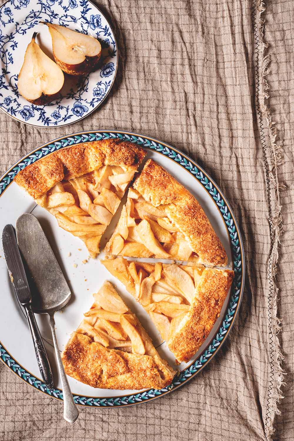 Galette de peras francesa Se ve un mantel gris vintage. En el mantel hay un plato blanco dónde está la galette de peras francesa cortada. En el plato hay una pala de postres pequeña. Al lado del plato blanco hay un plato pequeño, con dibujos azules, con peras cortadas en la mitad.