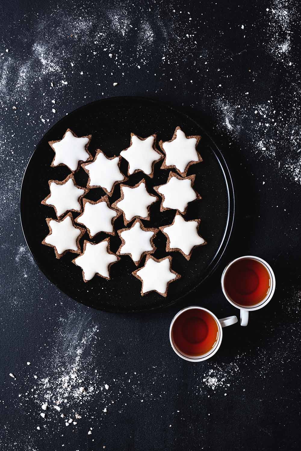 En una mesa negra hay un plato negro lleno de Zimtsterne. Al lado del plato hay dos tazas de té recién hecho. En la mesa se ven migas de azúcar.