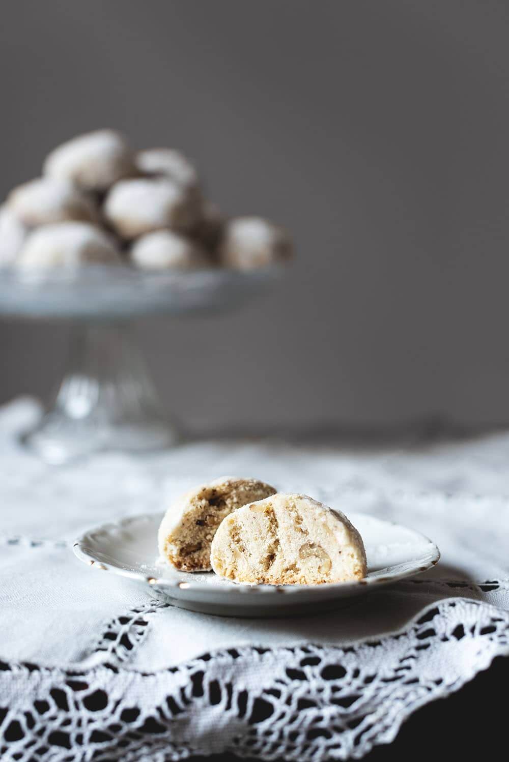 Galletas de té rusas crujientes En una mesa de madera hay un mantel blanco. Encima del mantel hay un porta tartas llenos de galletas de té rusas. Delante de este porta tartas hay un plato pequeño con una Galleta de té rusa cortada en la mitad. Está crujiente y se ven unas nueces también.