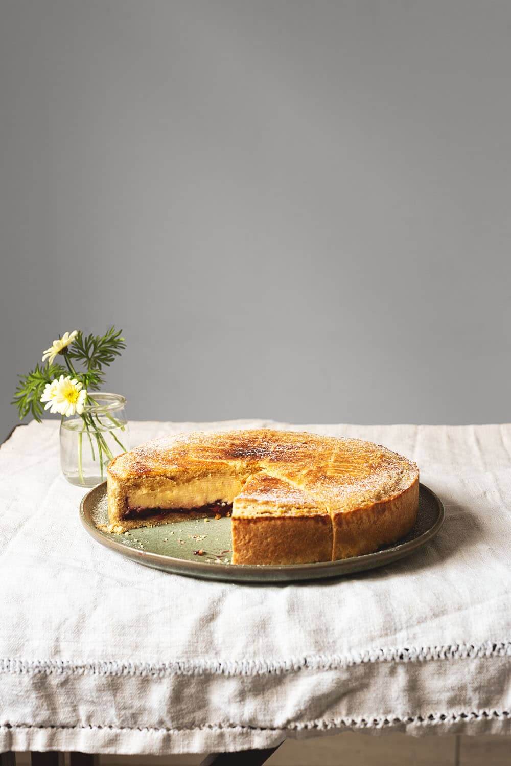 Se ve un mantel blanco tradicional. En el mantel hay un plato verde dónde está Gateau Basque ya cortado.Al lado hay un vaso pequeño con flores amarillas. Se ven las capas del postre. Crema pastelera, mermelada de cereza y masa quebrada.