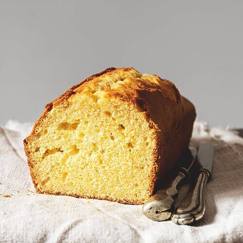En una mesa de madera hay un mantel blanco. Encima del mantel está el Gateau weekend ya cortado. Se ve muy esponjoso y tierno. Al lado del bizcocho hay dos cuchillos.