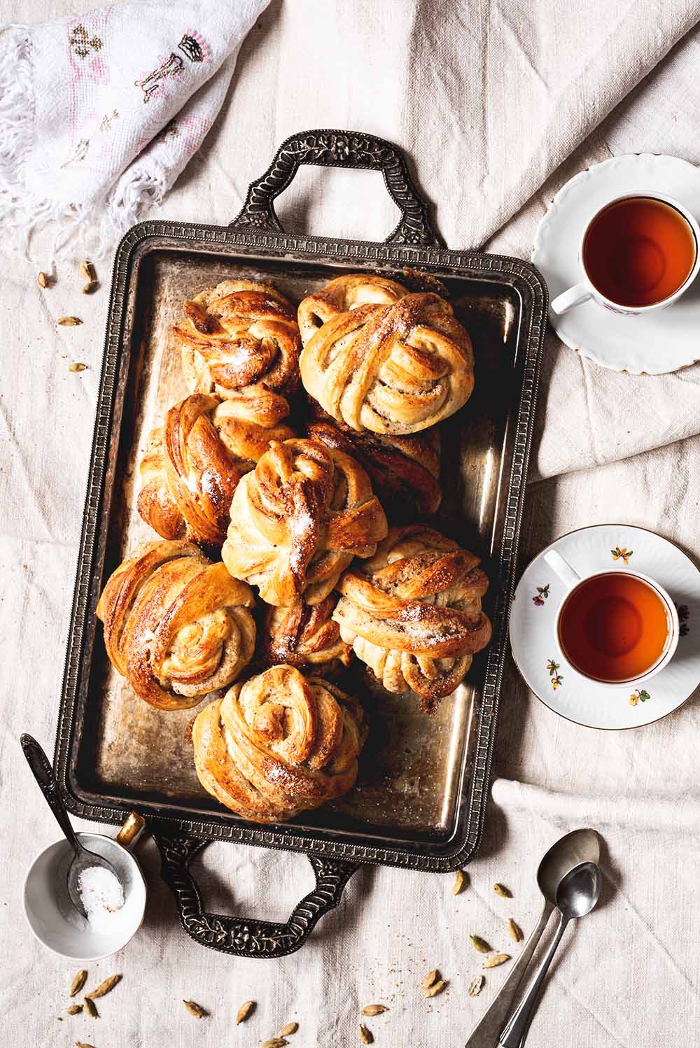 Kardemummabullar En una mesa está un mantel vintage. En el mantel hay una bandeja vintage, y encima muchos bollos cardamomo recién hechos. Alrededor de la bandeja hay dos tazas de té y un poco de azúcar , cucharas y cardamomo.
