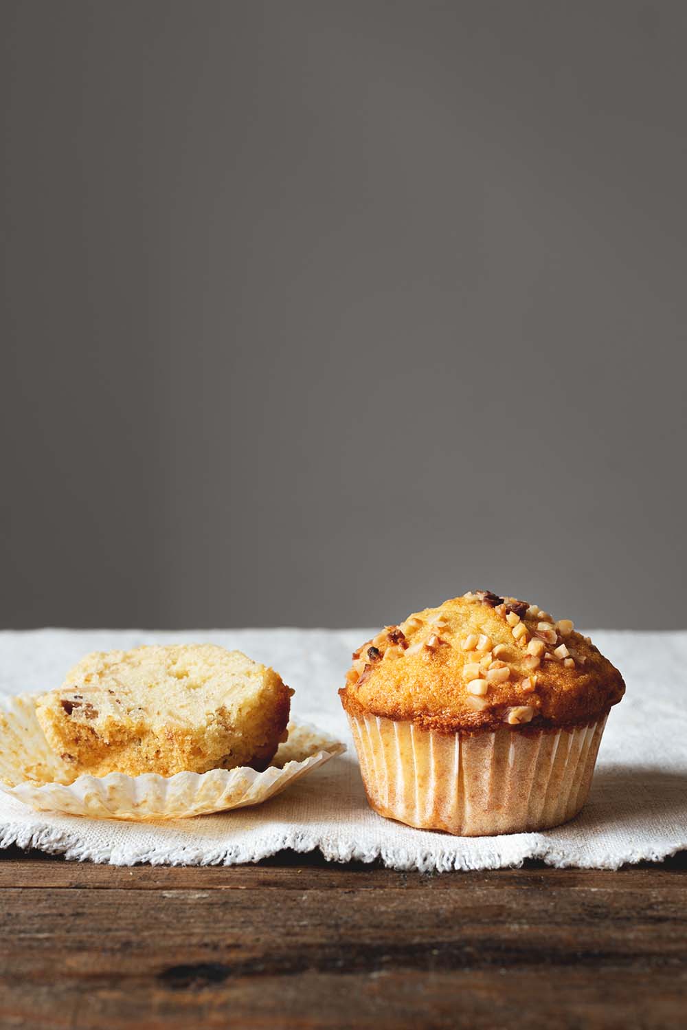 En una mesa de madera hay un mantel blanco. Encima del mantel hay dos magdalenas de almendra. Una de ellas está contada. Es muy esponjosa.
