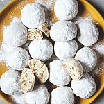 En una mesa gris hay un mantel amarillo. En el plato hay muchas galletas de té rusas. Se ven bonitas y cubiertas con azúcar glas.