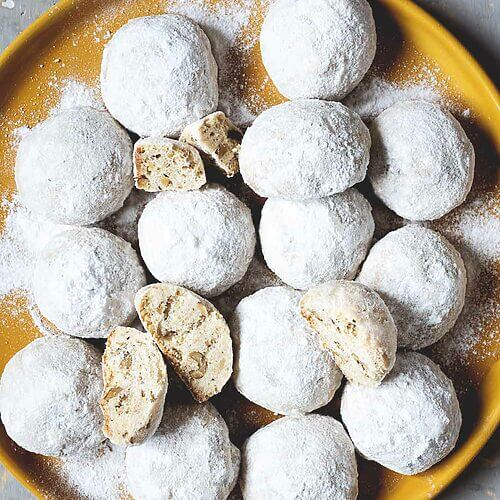 En una mesa gris hay un mantel amarillo. En el plato hay muchas galletas de té rusas. Se ven bonitas y cubiertas con azúcar glas.