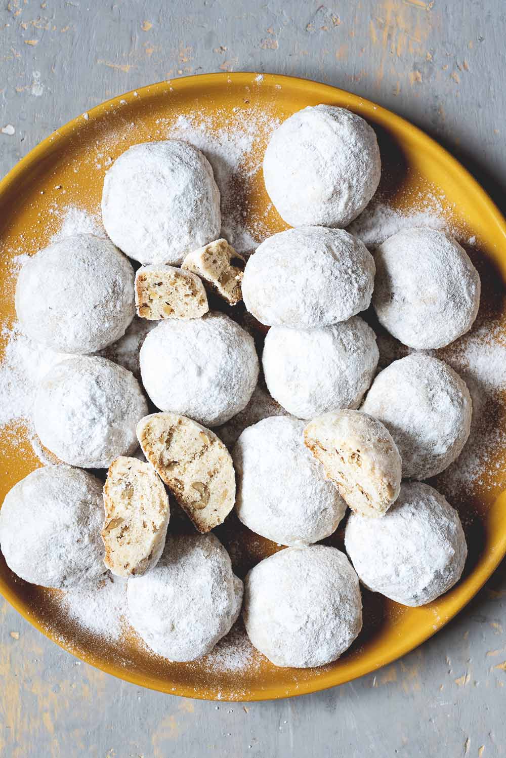 En una mesa gris hay un mantel amarillo. En el plato hay muchas galletas de té rusas. Se ven bonitas y cubiertas con azúcar glas.