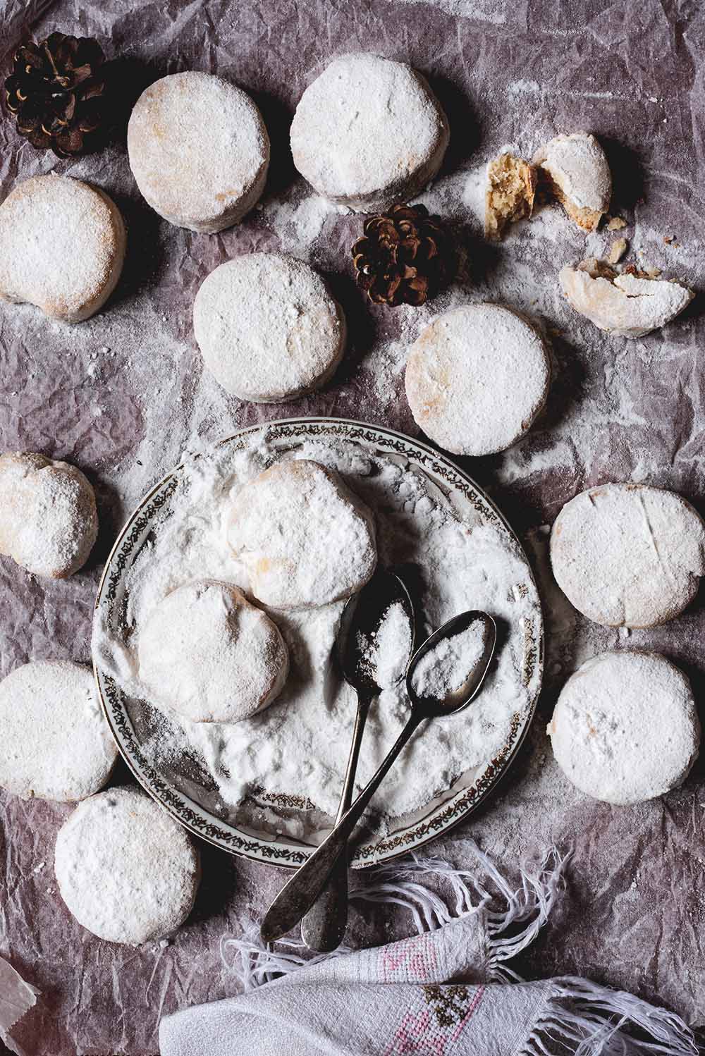 En una mesa hay papel de horno. En el papel están los Nevaditos de la abuela rebozados en azúcar glas. En el centro hay un plato lleno de azúcar glas , dos nevaditos recién rebozados y dos cucharas.