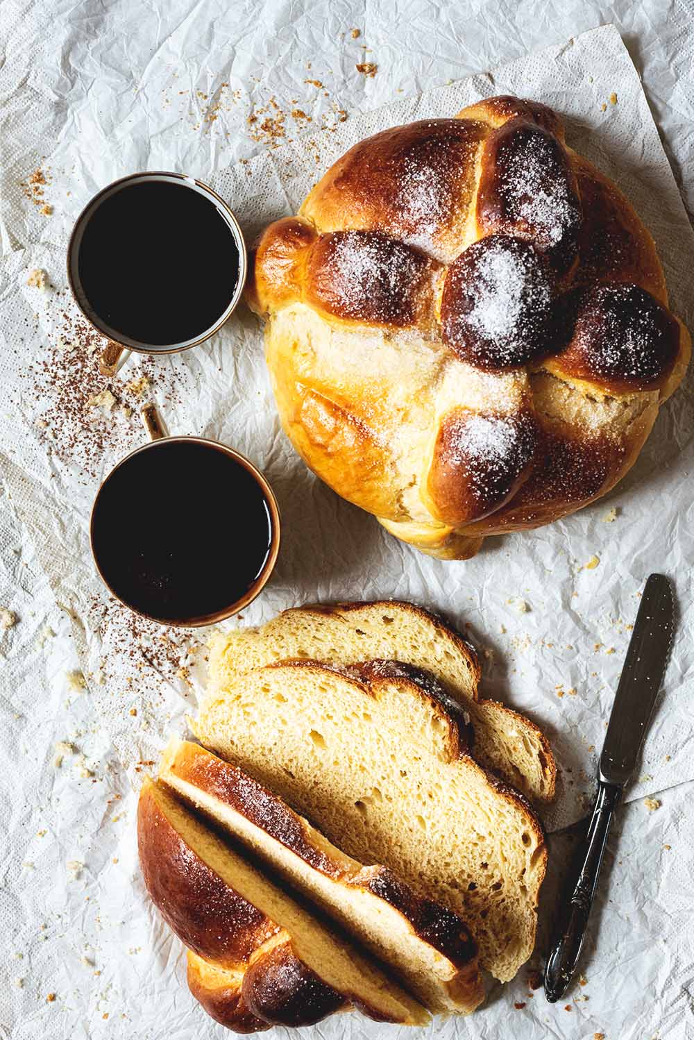 Pan de muerto mexicano En una servita hay dos panes de muerto. Uno ya está cortado. Se ve muy esponjoso. Al lado de los panes hay dos tazas de chocolate.