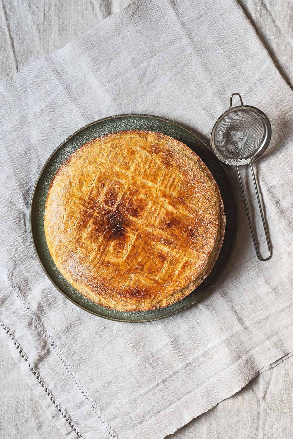 En el mantel hay un plato verde dónde está Pastel vasco casero. Se ven al lado del postre un colador pequeño con azúcar glas.