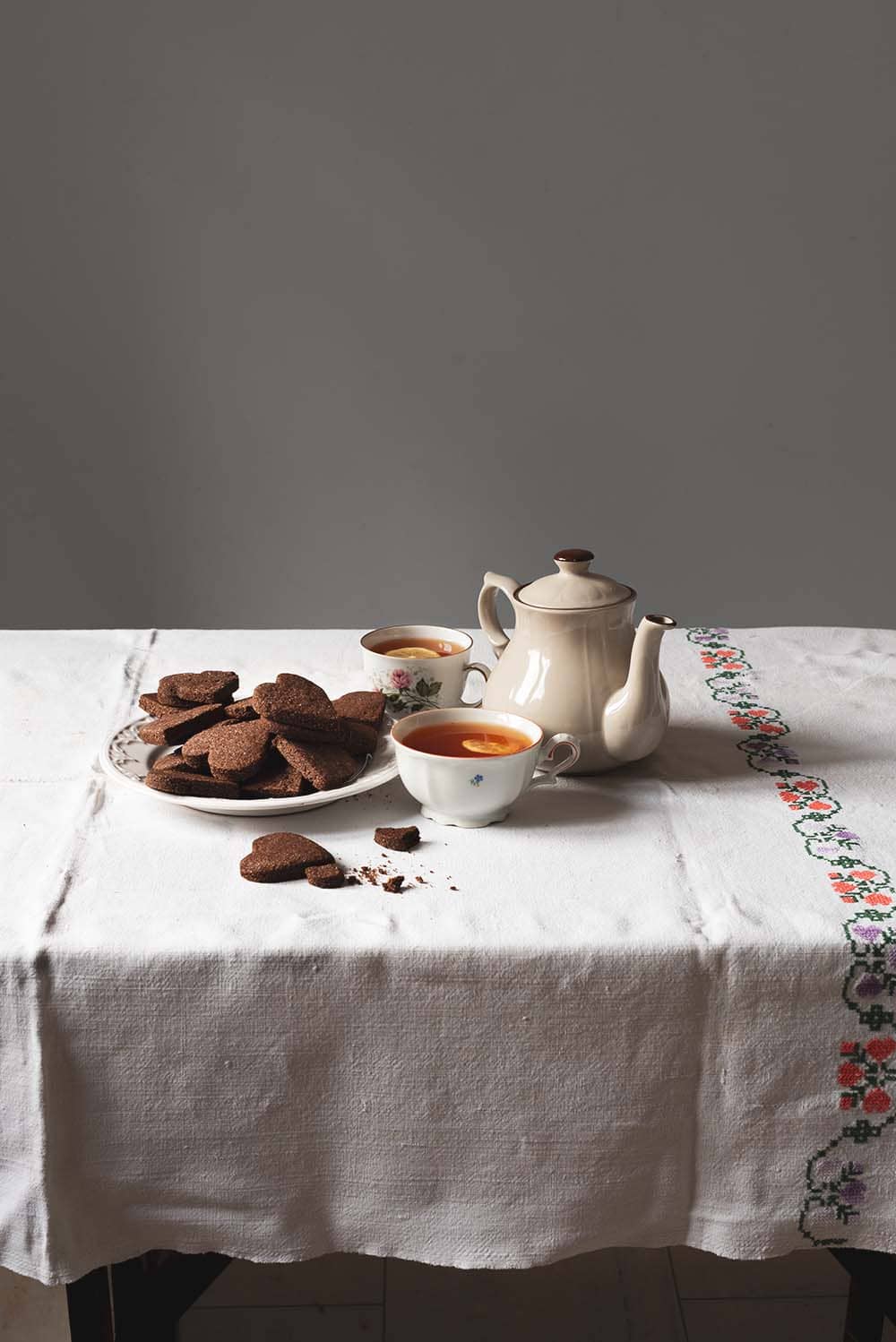 Receta de Basler Brunsli En una mesa hay un mantel blanco vintage. Encima hay un plato blanco lleno de Basler Brunsli. Alrededor hay una tetera y dos vasos de té recién hecho. También hay en la mesa migas de galleta. Se ven crujientes y con mucho sabor de chocolate.