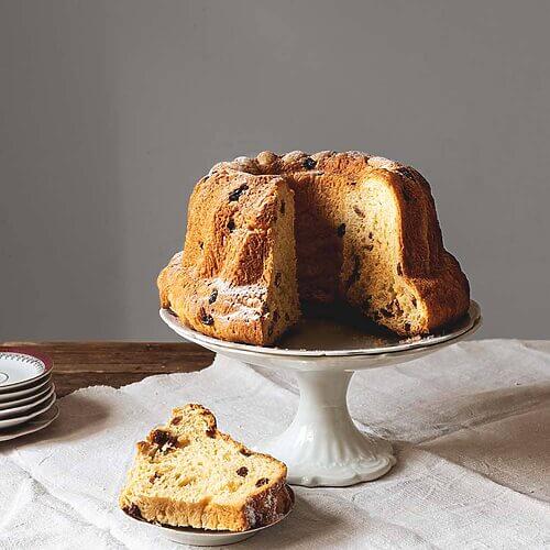 En una mesa de madera hay un mantel blanco. Encima del mantel hay un porta tartas. Encima está el Kouglof ya cortada. Se ve muy esponjoso y tierno. Delante del porta tartas hay un plato pequeño con una ración de Kouglof.