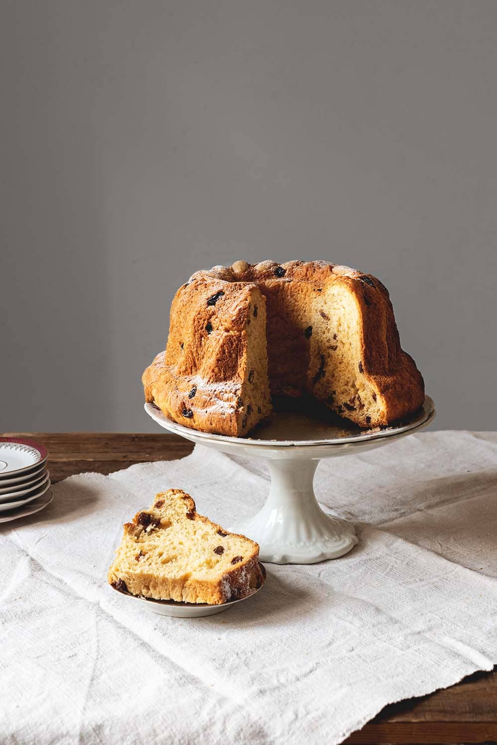 Receta de Kouglof de Alsacia En una mesa de madera hay un mantel blanco. Encima del mantel hay un porta tartas. Encima está el Kouglof ya cortada. Se ve muy esponjoso y tierno. Delante del porta tartas hay un plato pequeño con una ración de Kouglof.