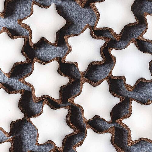 En una mesa hay papel de horno. En el papel están los Zimtsterne. Se ven muy bonitos su superficie blanca.