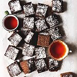 En mesa gris hay un papel de horno. Encima de este papel están los bizcochos de chocolate y coco ya cortados en cuadraditos. También se ven dos tazas de té recién hechos.