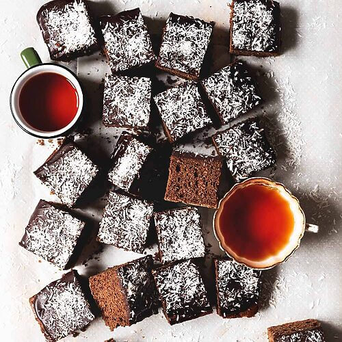 En mesa gris hay un papel de horno. Encima de este papel están los bizcochos de chocolate y coco ya cortados en cuadraditos. También se ven dos tazas de té recién hechos.