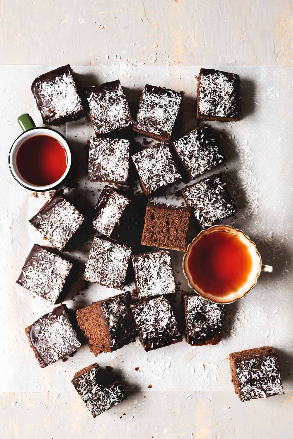 En mesa gris hay un papel de horno. Encima de este papel están los bizcochos de chocolate y coco ya cortados en cuadraditos. También se ven dos tazas de té recién hechos.
