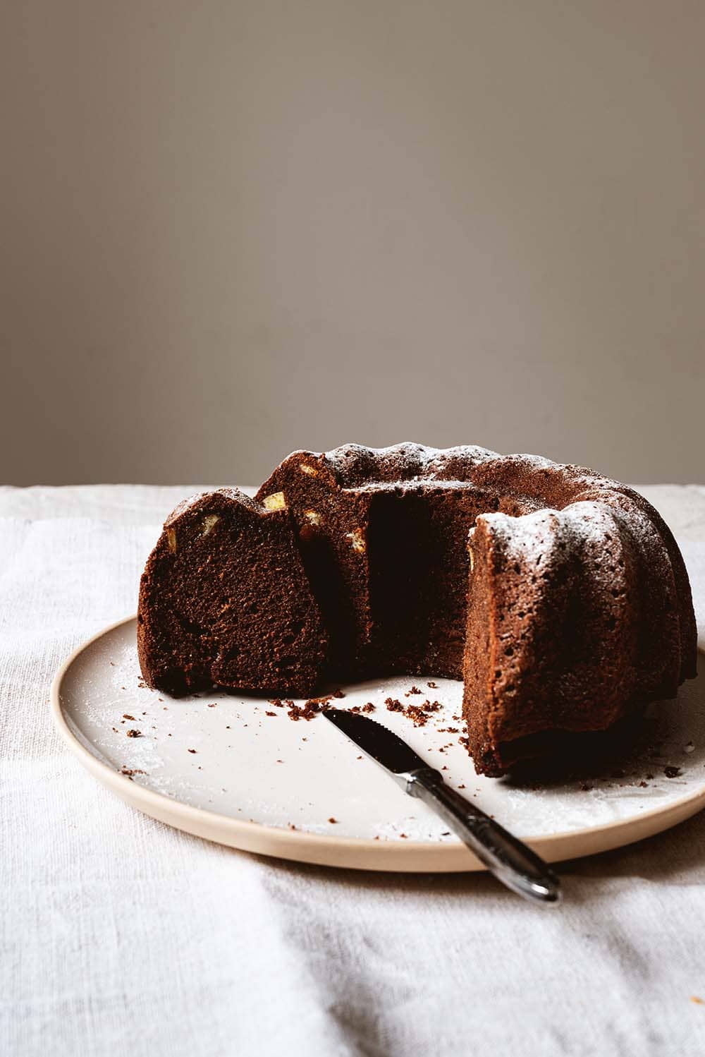 Receta de bizcocho de chocolate y naranja confitada Bizcocho de chocolate y naranja tierno y jugoso: En una mesa está un mantel vintage. En el mantel hay un plato blanco. Encima hay el bizcocho de chocolate con naranja ya está cortado. Se ve desde dentro muy jugoso y esponjoso.