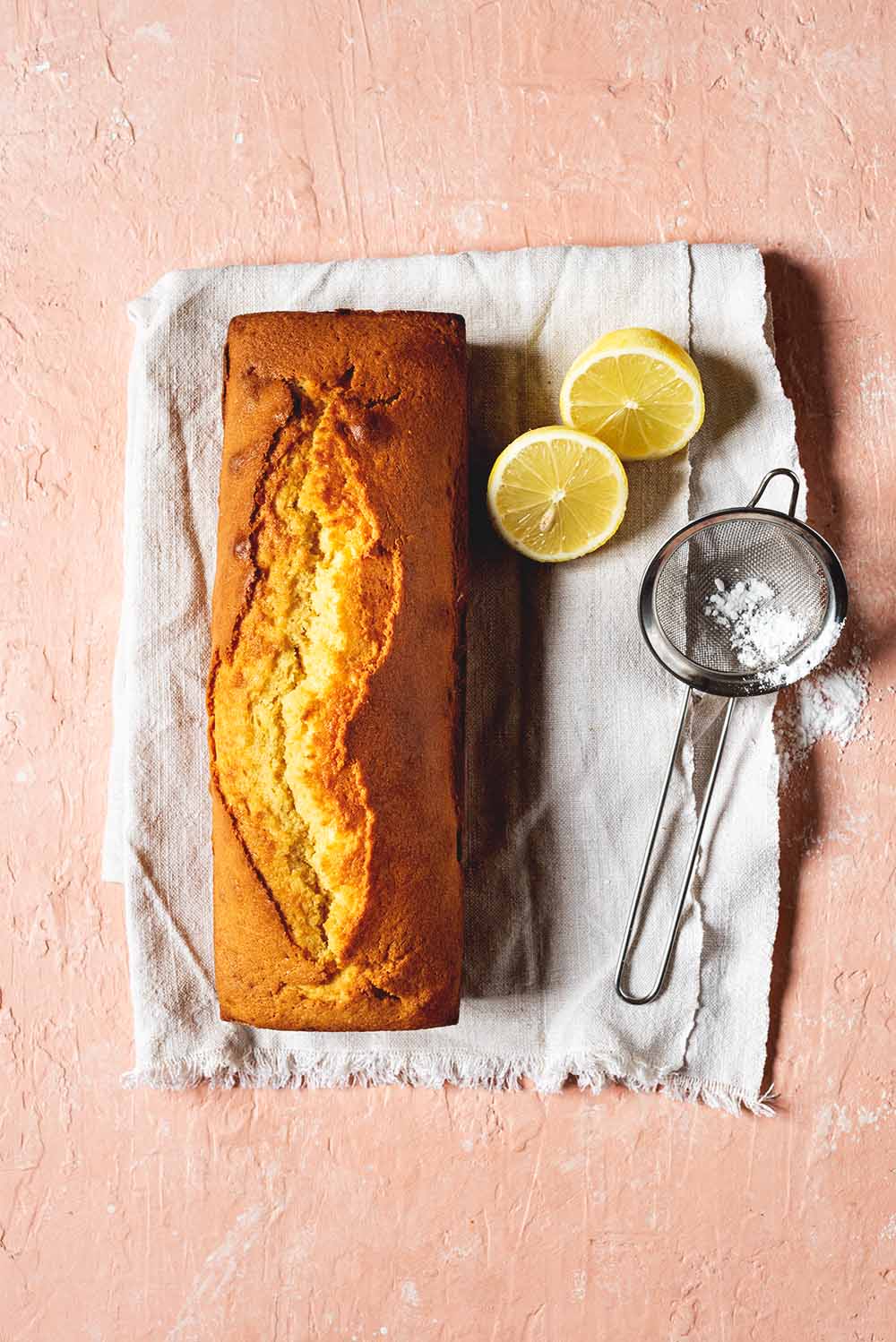 En una mesa de color rosa hay un mantel blanco. Encima del mantel está el Gateau weekend sin cortar. Al lado del bizcocho hay un limón cortado en la mitad y un colador pequeño con azúcar glas.