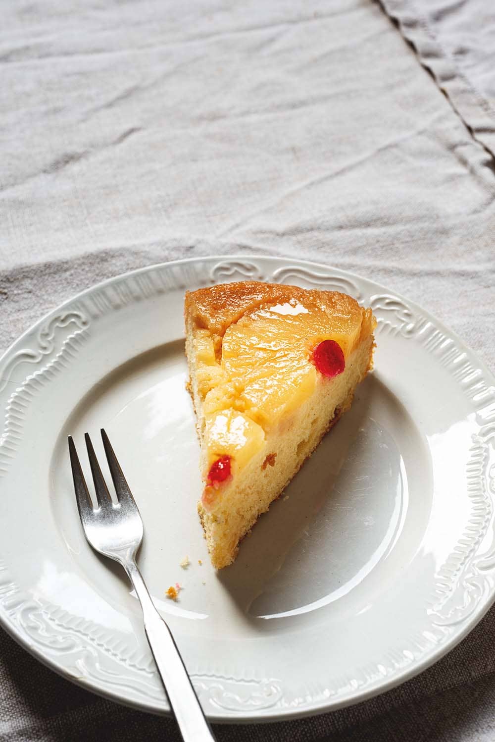 Receta de bizcocho de piña invertida En una mesa hay un mantel blanco Encima hay un plato blanco. En el plato hay una ración de bizcocho de piña invertido. Se ve muy jugoso y esponjoso. Al lado de la tarta hay un tenedor.