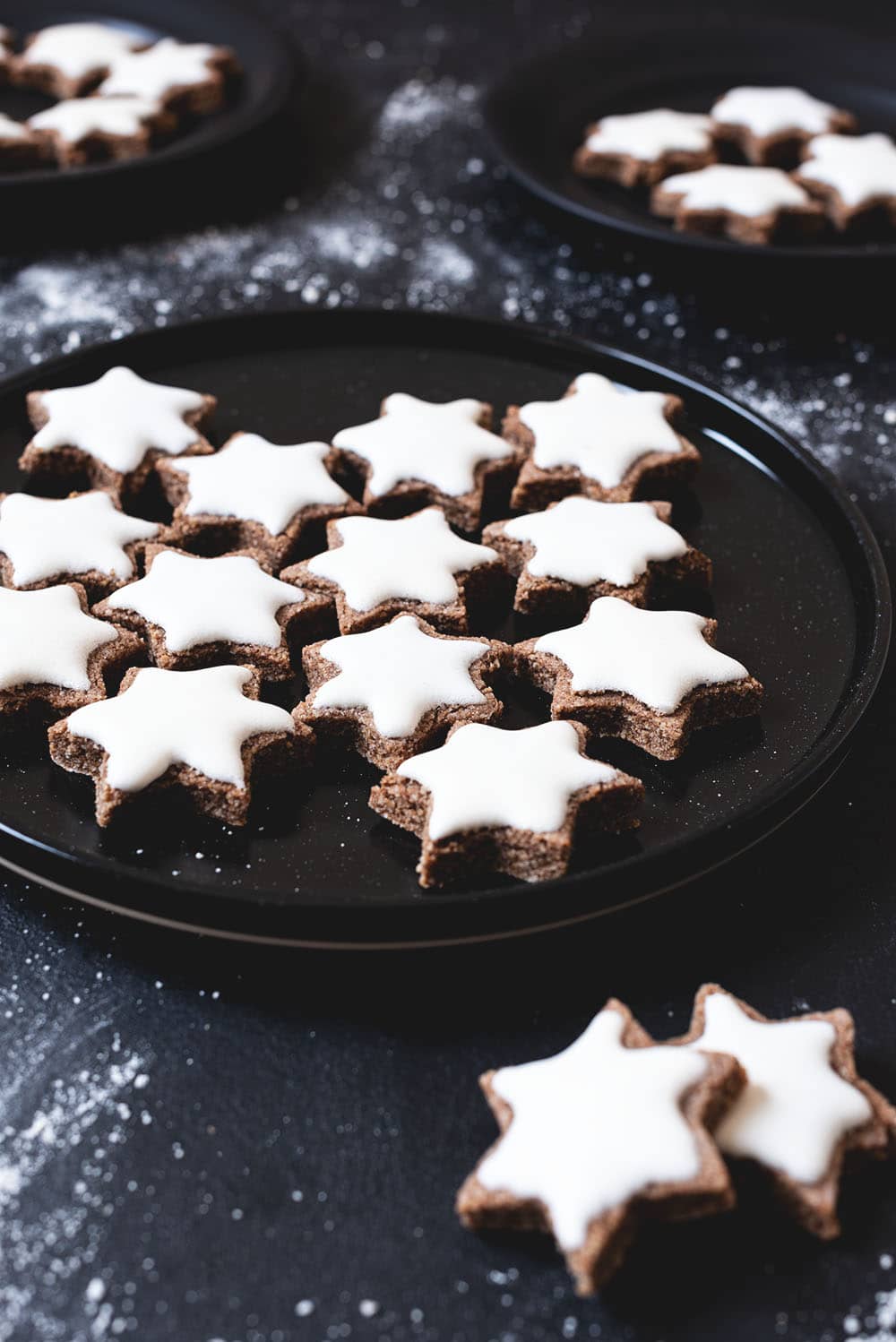 En una mesa negra hay tres platos negros. Cada una está llena de Zimtsterne. Se ven muy bonitos. La capa de azúcar blanca es muy suculenta. También se ven migas de azúcar en la mesa.