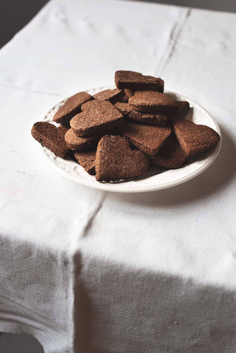 Receta de galletas suizas de chocolate En una mesa hay un mantel blanco vintage. Encima hay un plato blanco lleno de Basler Brunsli. Alrededor hay una tetera y dos vasos de té recién hecho. También hay en la mesa migas de galleta. Se ven crujientes y con mucho sabor de chocolate.