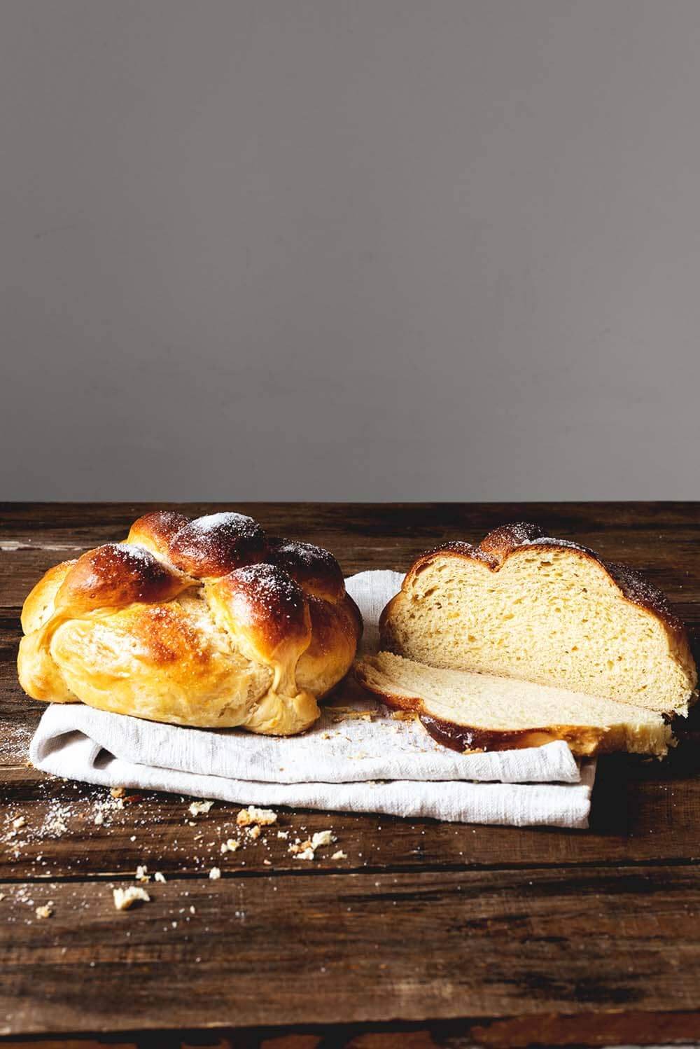 Receta de pan de muerto original en una mesa de madera hay paño de cocina blanca. Encima hay dos panes de muerto. Uno está cortado. Es muy esponjoso y tierno.