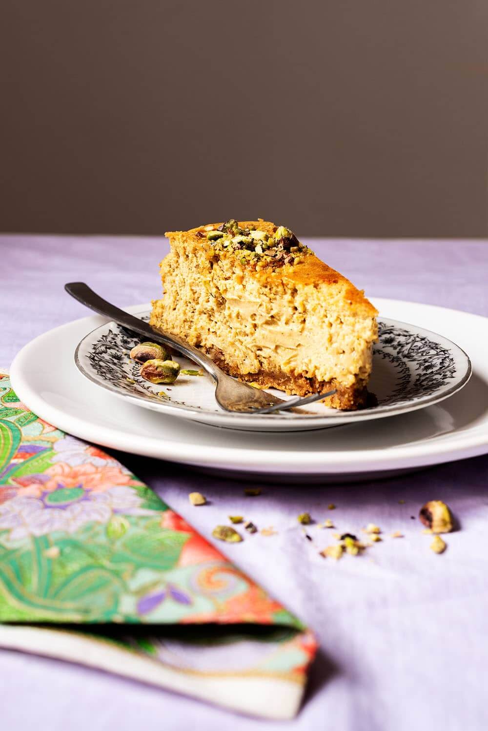 En una mesa hay un mantel de color lila. Encima hay dos platos encima una y otra. En el plato de arriba está una ración de tarta de queso de pistacho al horno junto con un tenedor. Se ve muy cremosa y rica. Al lado de los platos hay una servilleta vintage y migas de pistachos.