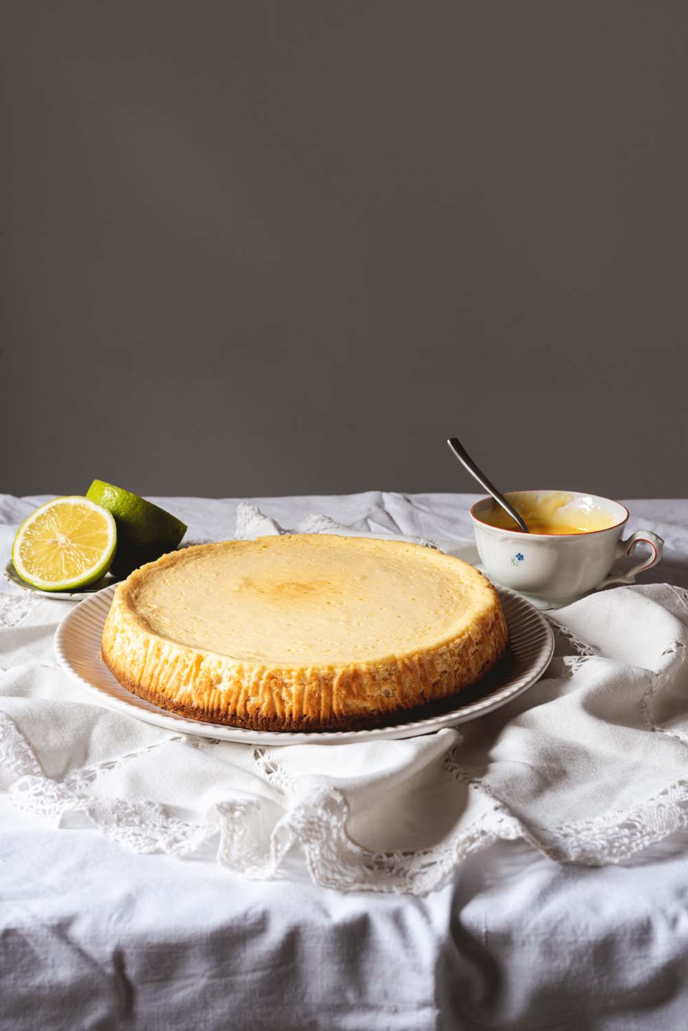 Receta de tarta de queso y limón al horno En una mesa hay un mantel blanco. En el mantel está un plato blanco. En el plato está la tarta de queso y limón al horno. Se ve suculenta. Al lado hay una taza con Lemon curd y limones cortados en la mitad.
