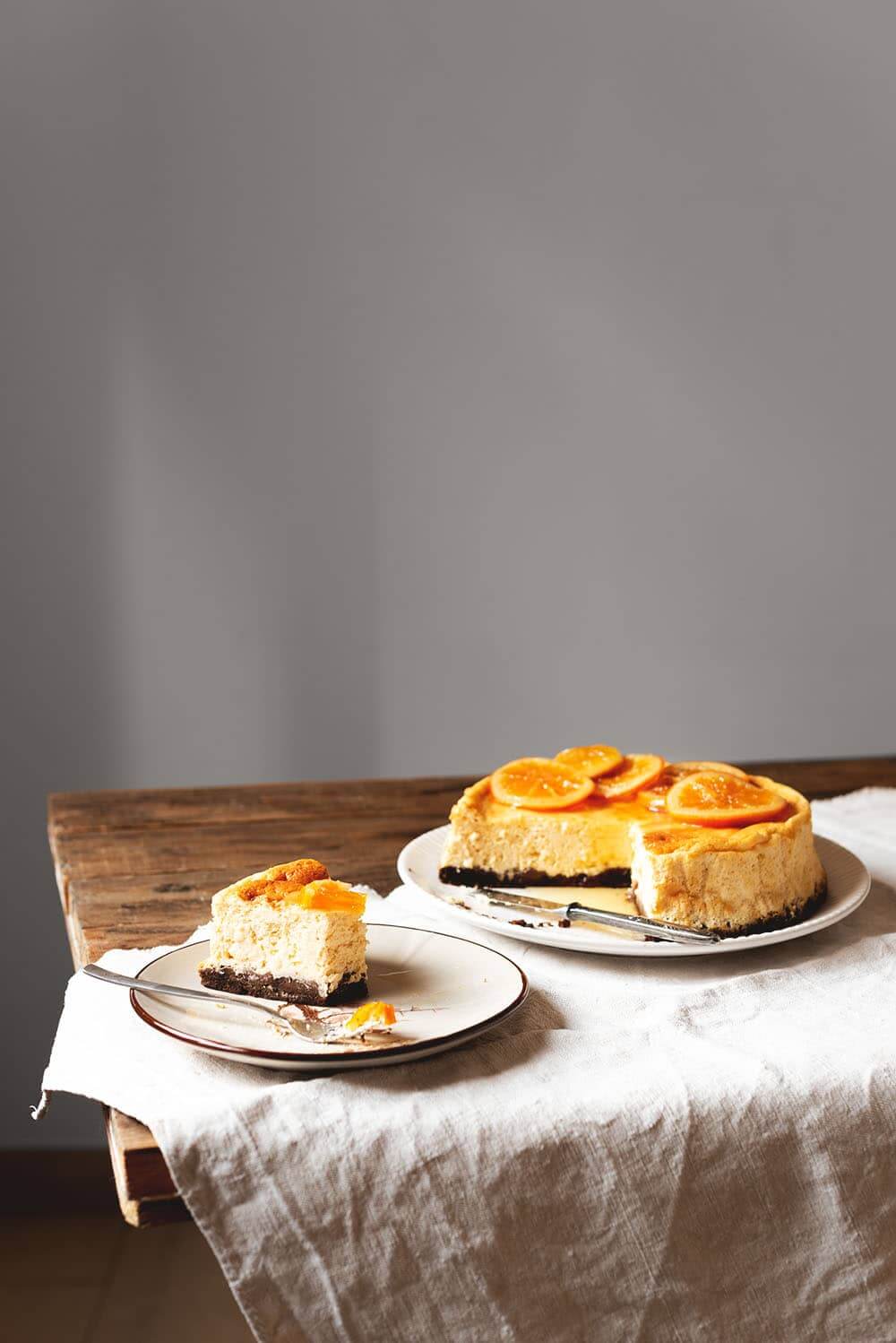 En una mesa hay un mantel blanco Encima hay un plato blanco. Encima está la de tarta de queso y naranja al horno ya cortadas. Encima de la tarta hay naranjas confitadas. Delante de este plato hay un plato más pequeño con una ración de tarta de queso y naranja junto con un tenedor.