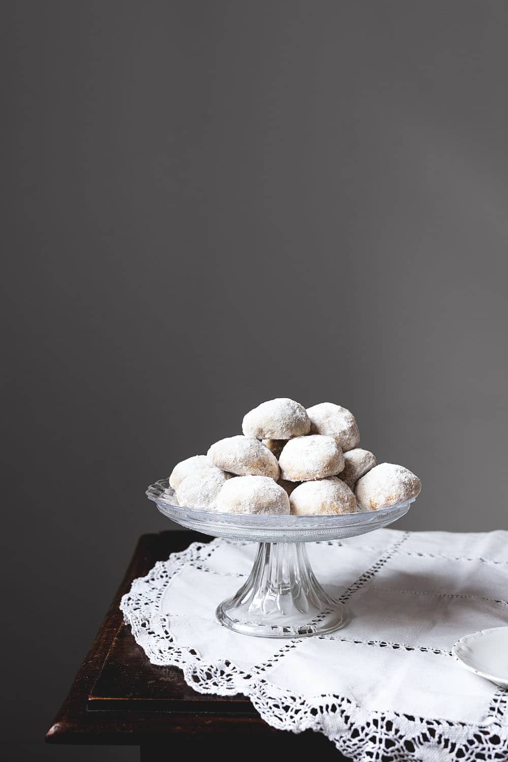 Russian tea cakes En una mesa de madera hay un mantel blanco vintage. Encima del mantel hay una porta tartas llenos de galletas de té rusas. Se ven bonitas y cubiertas con azúcar glas. Al lado de la porta tarta hay varios platos blancos pequeños.