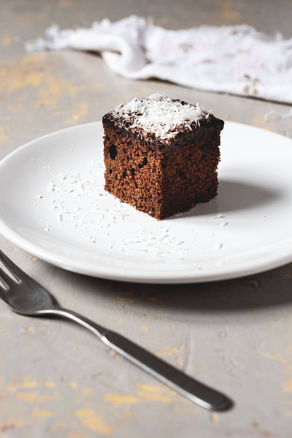 Skúffukaka En una mesa gris hay un plato blanco. En el plato está una ración de bizcocho de chocolate y coco. Se ve esponjoso y se ve la capa a de crema chocolate y el coco rallado encima de la crema. Al lado del plato hay un tenedor y una servilleta de algodón vintage.