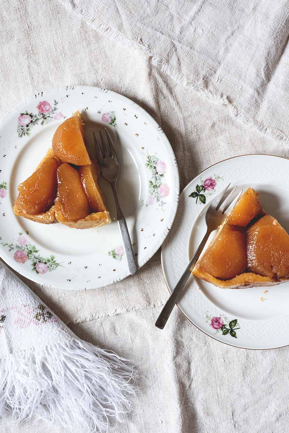 Se ve un mantel blanco tradicional. En el mantel hay dos platos pequeños. En cada una de los platos hay una ración de tarta Tatin de manzana tradicional con masa quebrada y tenedor.