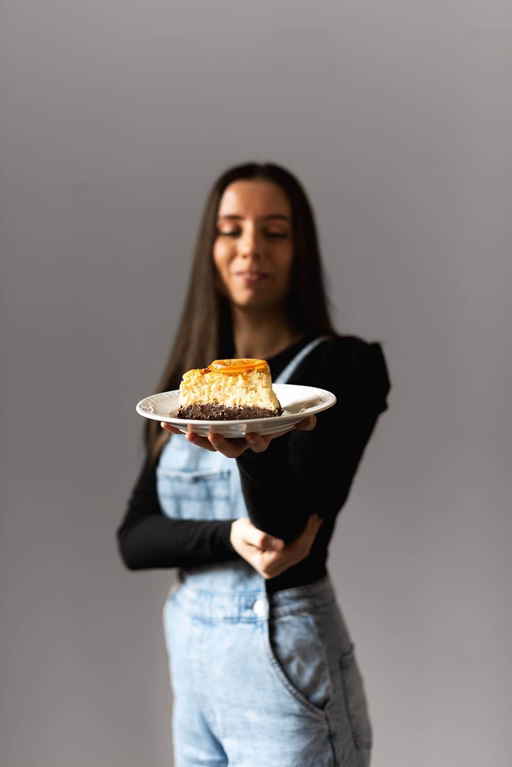 Se ve una persona con un plato en la mano. En este plato hay una ración de tarta de queso y naranja al horno. Se ve la tarta cremosa y jugosa.