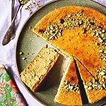 En una mesa hay un mantel de color lila. Encima hay un alto verde. Encima esta tarta de queso de pistacho al horno ya cortada. Se ve muy cremosa. Al lado del plato verde hay cucharas, una servilleta vintage y platos pequeños.