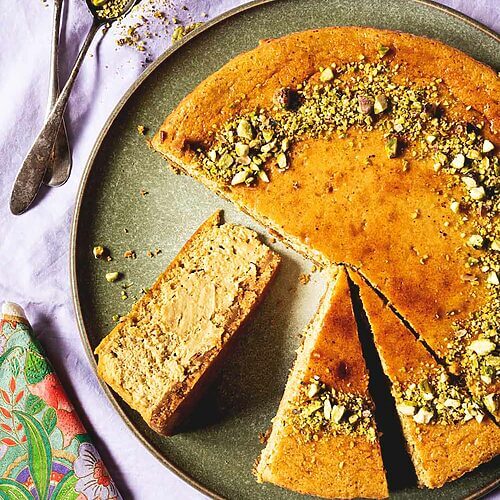 En una mesa hay un mantel de color lila. Encima hay un alto verde. Encima esta tarta de queso de pistacho al horno ya cortada. Se ve muy cremosa. Al lado del plato verde hay cucharas, una servilleta vintage y platos pequeños.