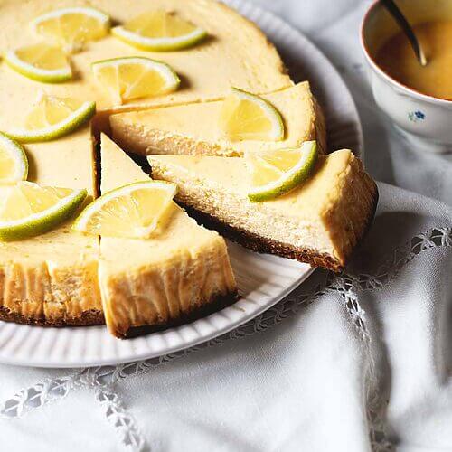 En la mesa cubierta con mantel blanco está la tarta de queso y limón al horno. Se ve muy cremosa. Encima hay para decorar rodajas de limones. Al lado del plato hay una hay en una taza con Lemon curd.