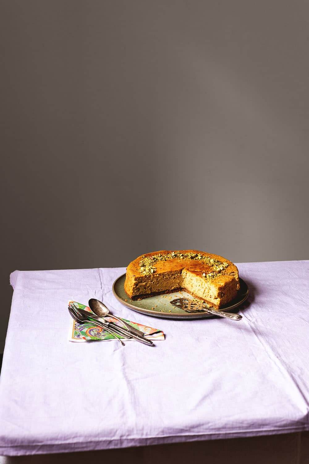 En una mesa hay un mantel de color lila. Encima hay un plato verde. Encima esta tarta de queso de pistacho al horno ya cortada junto con una pala de tartas vintage. Al lado del plato hay una servilleta vintage y cucharas y tenedores.