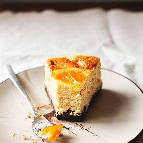 En una mesa hay un mantel blanco Encima hay un plato blanco. Encima está una ración de tarta de queso y naranja al horno junto con un tenedor. Se ve la tarde muy cremosa.