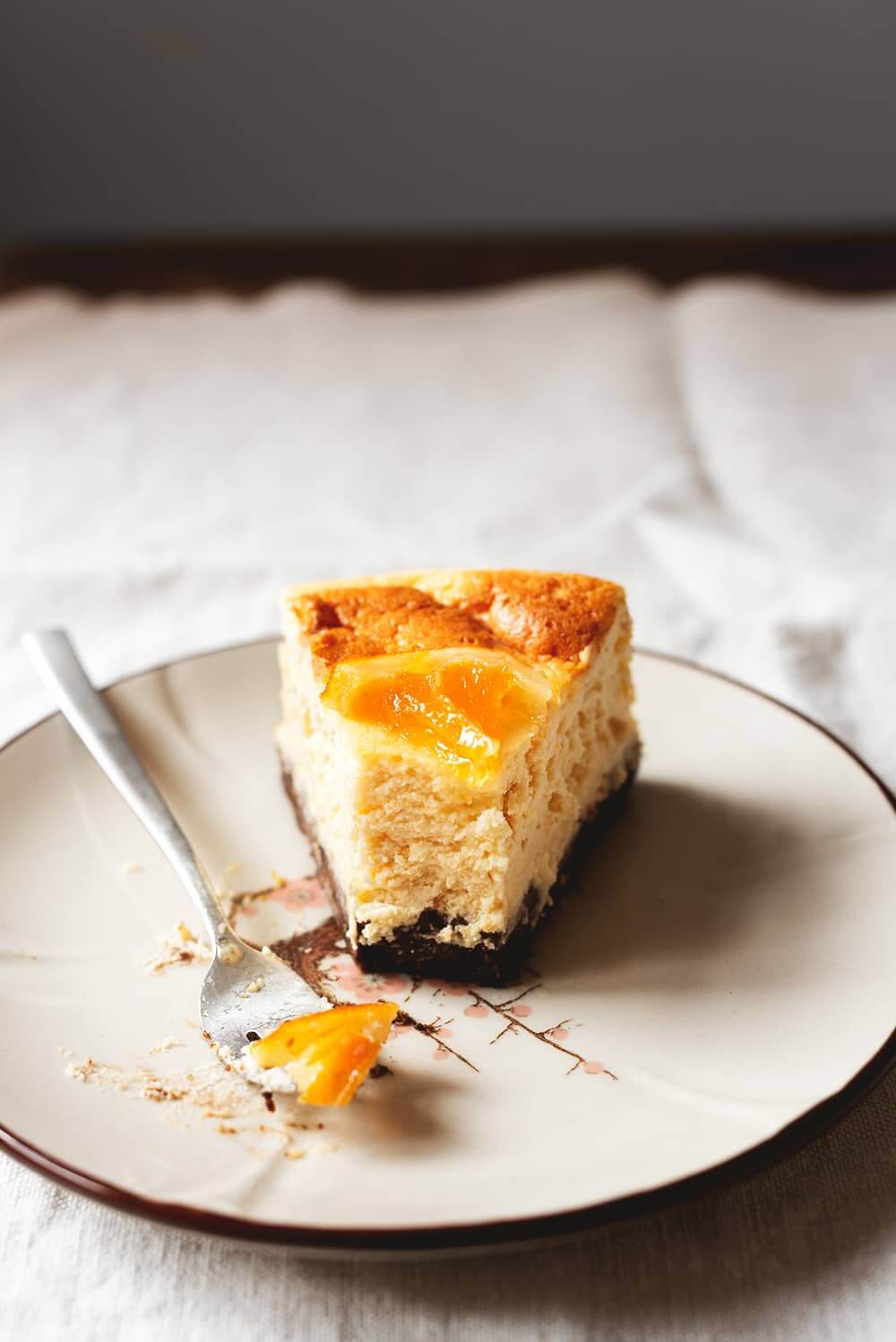 En una mesa hay un mantel blanco Encima hay un plato blanco. Encima está una ración de tarta de queso y naranja al horno junto con un tenedor. Se ve la tarde muy cremosa.