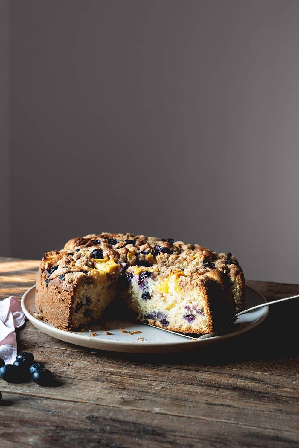 Bizcocho americano de melocotón y arándanos En una mesa de madera hay un plato blanco. Encima está el bizcocho americano de melocotón y arándanos ya cortado. Se ve muy jugoso y esponjoso. En el plato hay una pala de tarta y alrededor del plato hay una servilleta de tela rosa y arándanos frescos.