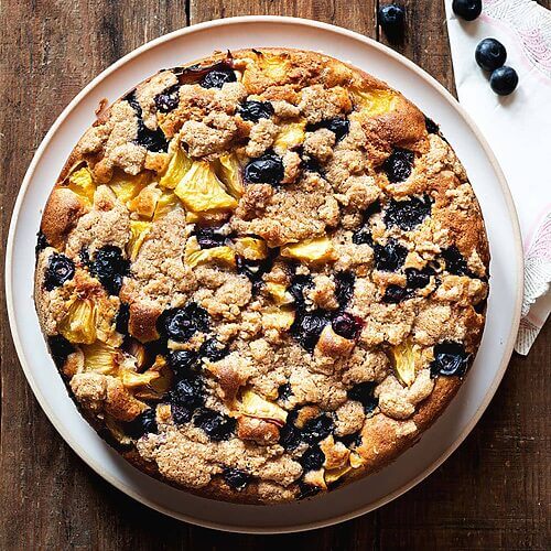 Es una foto cenital, donde se ve que el postre está en una mesa de madera. No está cortado todavía. Se ve la superficie del bizcocho con crumble, melocotón y arándanos: crumble dulce y crujiente y frutas. Al lado del postre hay una servita de tela rosa y arándanos frescos.