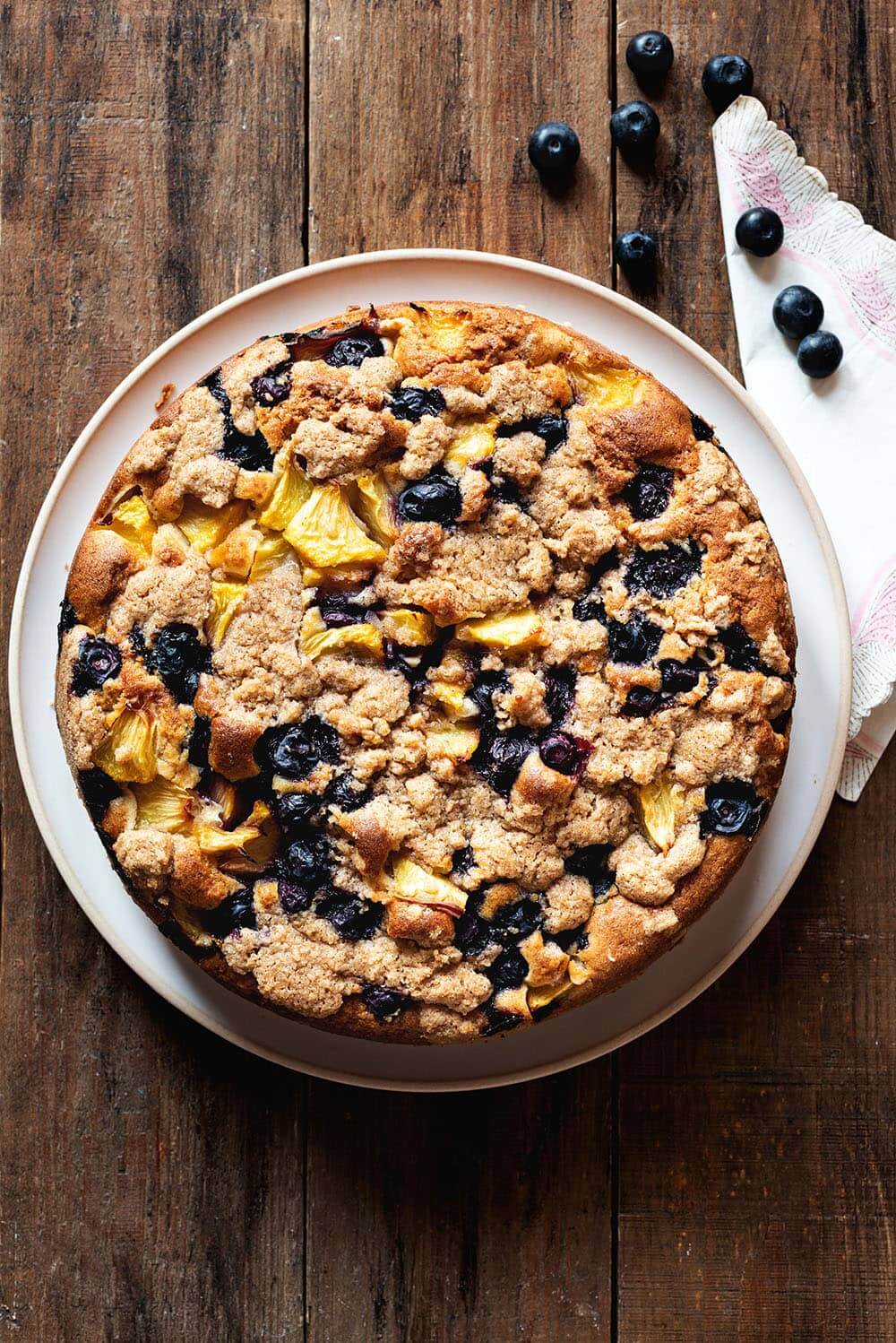 Es una foto cenital, donde se ve que el postre está en una mesa de madera. No está cortado todavía. Se ve la superficie del bizcocho con crumble, melocotón y arándanos: crumble dulce y crujiente y frutas. Al lado del postre hay una servita de tela rosa y arándanos frescos.