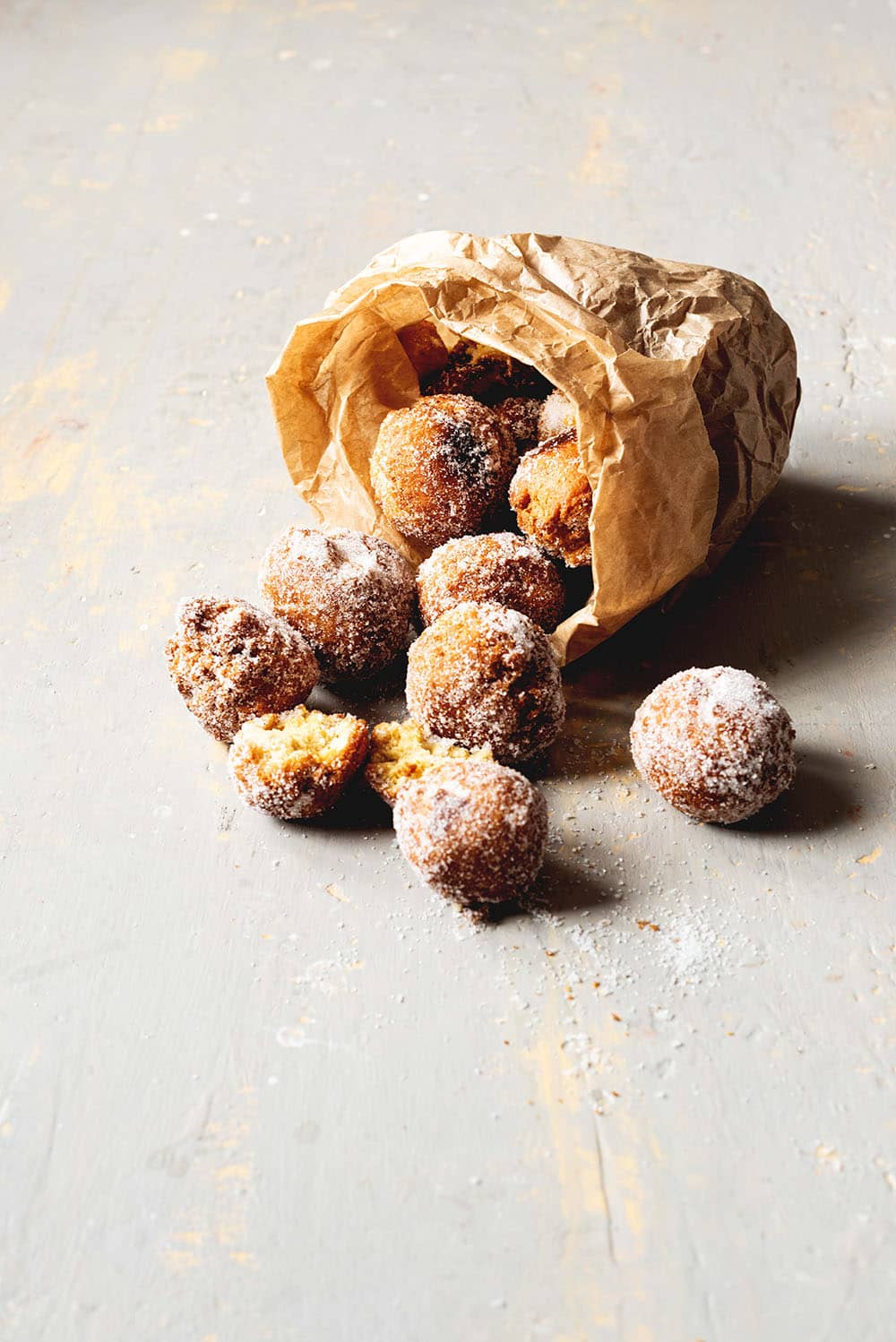 En una mesa gris hay una bolsa de papel lleno de buñuelos italianos para carnaval volcado. se ven deliciosos y esponjosos.