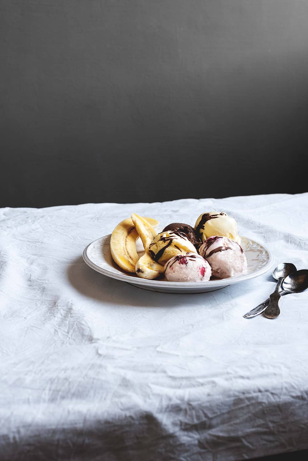 En una mesa hay un mantel blanco. Encima hay un plato blanco. Encima está el postre Banan Split. Hay al lado del plato hay dos cucharas soperas.